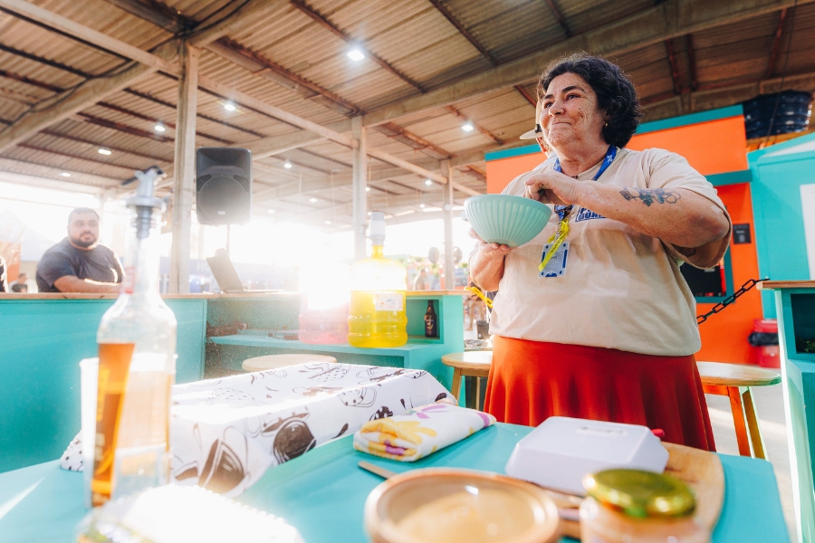 Nesta imagem, a mulher aparece em pé, segurando a tigela de cerâmica verde-água com as duas mãos à altura do peito, enquanto mexe o conteúdo com uma colher. Ela exibe um sorriso discreto e satisfeito, olhando para algo à sua frente fora do quadro. O plano é aberto, revelando mais do ambiente: um grande galpão com estrutura de telhado aparente e iluminação por refletores. À esquerda, em primeiro plano sobre a mesa azul, destaca-se uma garrafa de vidro com bico dosador e um pano de prato estampado. Ao fundo, um homem observa a cena sentado atrás do balcão, e a luz solar entra lateralmente, criando um efeito de brilho intenso (flare) que reforça a atmosfera vibrante e produtiva do evento.