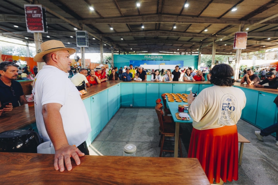 Uma mulher está de costas, no lado direito do quadro, posicionada à frente de uma longa mesa azul em formato de "U", onde várias pequenas tigelas de madeira estão organizadas para degustação. Ela veste a mesma saia vermelha e uma camiseta bege com a estampa "Orgulho do meu Rio Grande do Norte". À esquerda, o homem de chapéu de palha observa a cena apoiado em um balcão de madeira. Ao fundo, uma plateia atenta de diversas pessoas acompanha a apresentação, cercada pela estrutura industrial do galpão, que exibe faixas coloridas penduradas no teto e um painel ao fundo com a inscrição "FEAQUARN".