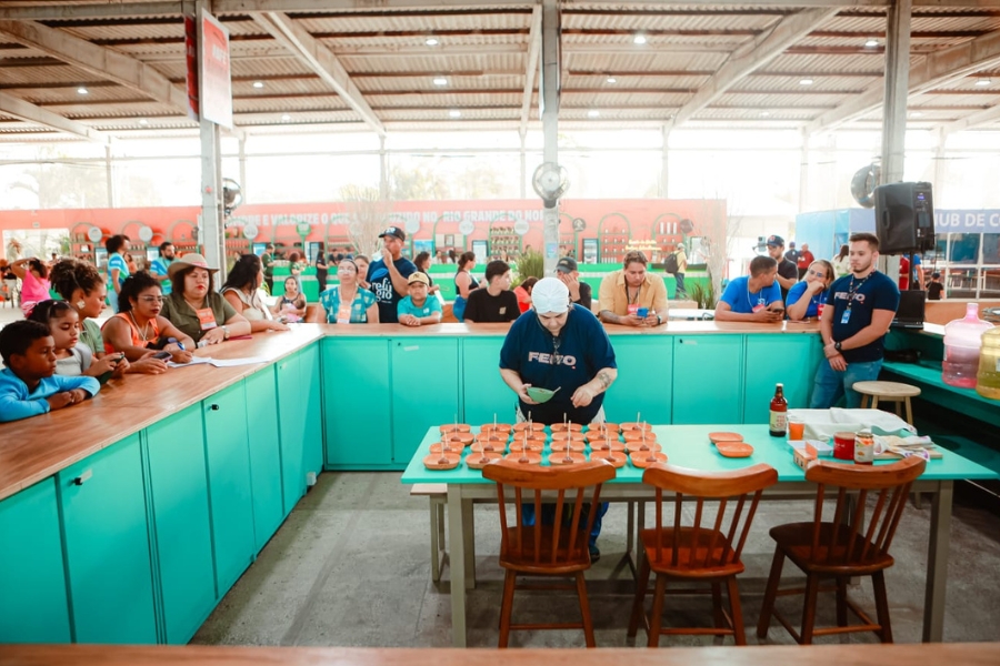 Uma mulher vestindo uma camiseta azul-marinho e turbante branco aparece centralizada atrás de uma mesa azul repleta de pequenas tigelas dispostas em fileiras organizadas para degustação. Ela está inclinada, focada em finalizar a montagem de cada porção com cuidado. Ao redor da estrutura em "U" formada por balcões azuis e tampos de madeira, diversas pessoas — adultos e crianças — observam atentamente o processo.