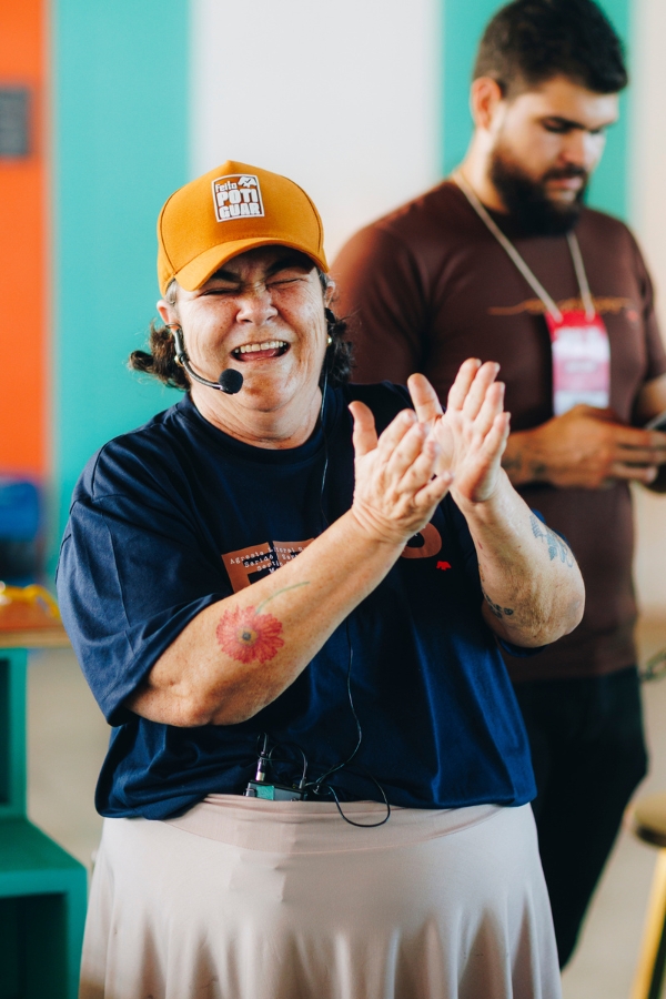Uma mulher aparece em um momento de pura celebração e alegria. Ela está de pé, com os olhos fechados em um sorriso largo, enquanto aplaude com entusiasmo. Ela usa uma camiseta azul-marinho, uma saia bege e um boné de cor ocre com a marca "Feito Potiguar". No antebraço direito, destaca-se uma tatuagem de flor avermelhada. Ela ainda utiliza o microfone auricular, indicando o encerramento ou um momento de clímax em sua apresentação. Ao fundo, um homem com barba e camiseta marrom observa a cena, e as cores vibrantes em tons de azul e laranja nas paredes reforçam a energia festiva do evento.