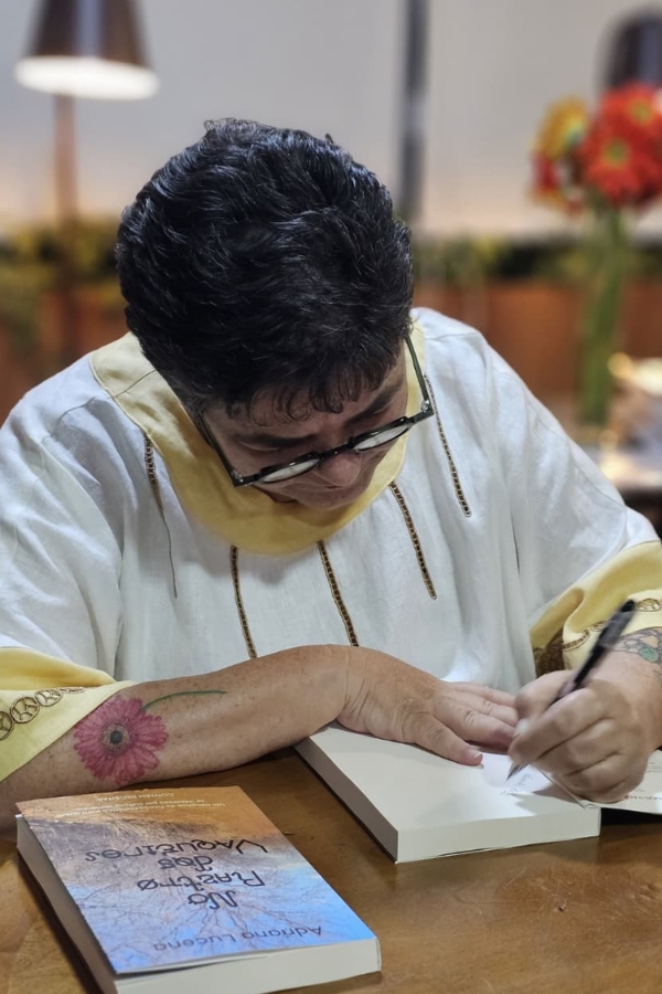 Uma mulher está sentada à mesa, inclinada sobre um exemplar aberto do seu livro, intitulado "No Rastro dos Vaqueiros", enquanto escreve uma dedicatória com uma caneta preta. Ela usa óculos de grau e uma túnica branca com detalhes em amarelo suave na gola e nas mangas. Sua tatuagem de flor avermelhada no antebraço direito ganha destaque sobre a mesa de madeira. Ao lado, um exemplar fechado do livro revela a capa com uma imagem de paisagem árida sob um céu azul. O ambiente ao fundo é acolhedor, com luzes quentes e um vaso de flores laranjas desfocado, sugerindo uma sessão de autógrafos ou um momento pessoal de registro.