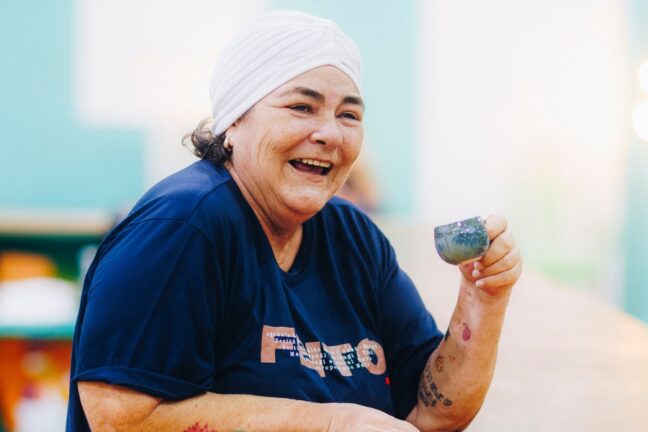 Uma mulher de pele clara e meia-idade sorri alegremente enquanto segura uma pequena xícara de café azulada. Ela usa um turbante branco de tecido drapeado, uma camiseta azul-marinho com estampas em letras grandes e possui algumas tatuagens visíveis no antebraço e pulso. O ambiente ao fundo está levemente desfocado, sugerindo um local iluminado com tons de azul e madeira, transmitindo uma sensação de acolhimento e descontração.