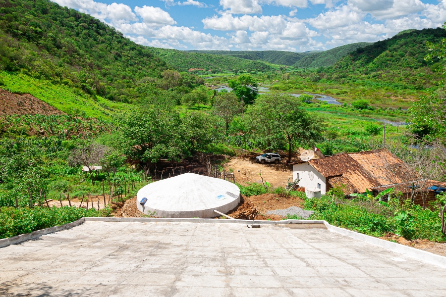 Esta fotografia em plano aberto e perspectiva aérea revela uma paisagem exuberante do semiárido brasileiro, destacando a convivência harmoniosa entre a infraestrutura hídrica e a natureza. No centro inferior, uma cisterna de placas circular com cúpula branca destaca-se no terreno, conectada a um amplo calçadão de cimento em declive no primeiro plano, projetado para captar a água da chuva. Ao lado, encontra-se uma casa de campo simples com telhado de barro, antena parabólica e um carro branco estacionado no quintal. A propriedade é cercada por uma densa vegetação verde que cobre morros ondulados ao fundo, onde um rio serpenteia pelo vale sob um céu azul vibrante, repleto de nuvens brancas e volumosas, simbolizando a segurança hídrica e a resistência da agricultura familiar.