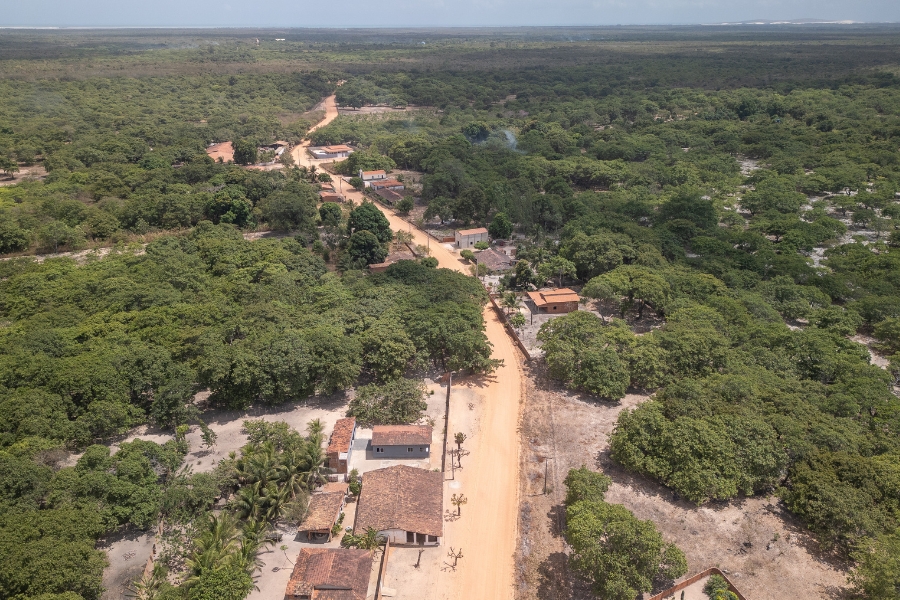 Esta fotografia aérea revela uma pequena comunidade rural inserida no coração de uma vasta área de vegetação nativa. Uma estrada de terra de tom alaranjado serpenteia pelo centro da imagem, servindo de eixo para algumas casas simples de telhado de barro espalhadas ao longo de suas margens. As construções são cercadas por árvores frondosas e quintais arenosos, com a mata densa e verde se estendendo até onde a vista alcança em todas as direções. Ao fundo, a linha do horizonte revela uma transição suave para áreas de dunas sob um céu claro