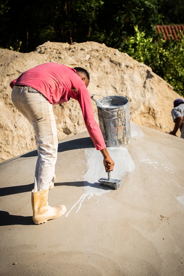 A imagem mostra um homem trabalhando no acabamento externo da cúpula de uma cisterna de placas. Ele está curvado para a frente, vestindo uma camiseta rosa de mangas compridas, calça de sarja clara e botas de borracha bege. Com a mão direita, ele utiliza uma trincha grossa para aplicar uma camada de cal branca sobre a superfície curva e cinzenta de cimento, visando a impermeabilização e a proteção térmica da estrutura. Ao lado dele, sobre a cúpula, há um balde metálico com resíduos da tinta branca. Ao fundo, vê-se um monte de areia clara, vegetação verde e o telhado de uma construção, indicando que a obra ocorre em um ambiente rural sob a luz forte do sol.