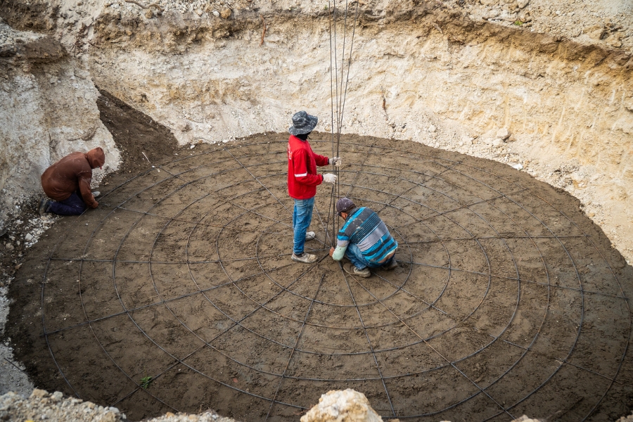 A imagem mostra a construção da base de uma cisterna em um terreno escavado de terra clara e arenosa. No centro de uma grande cavidade circular, três homens trabalham na montagem de uma estrutura de ferro: dois deles estão agachados e um está de pé, vestindo uma camisa vermelha de mangas compridas e chapéu de abas largas, segurando vergalhões verticais. A base no chão é formada por uma grade metálica em formato radial e concêntrico, lembrando uma teia de aranha gigante, sobre a terra batida e nivelada. A cena, capturada de um ângulo superior, evidencia o trabalho manual e técnico envolvido na infraestrutura hídrica em áreas rurais.