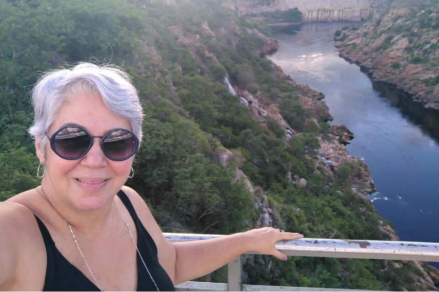 Selfie de uma mulher branca, com cabelos curtos e grisalhos que sorri suavemente para a câmera usando óculos de sol redondos de armação escura, brincos de argola prateados e uma regata preta. Ela está em um mirante, com a mão esquerda apoiada em um corrimão metálico cinza. O plano de fundo revela uma vista panorâmica impressionante de um cânion profundo, por onde corre um rio em tons de azul escuro. As encostas são íngremes e cobertas por vegetação verde densa, e ao fundo, é possível avistar a estrutura de uma usina hidrelétrica cruzando o rio sob a luz suave do entardecer