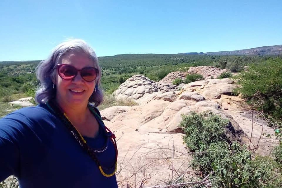Selfie de mulher. Em primeiro plano, à esquerda, ela sorri para a câmera. Trata-se de uma mulher branca com cabelos curtos e ondulados em tons de grisalho e azul-violeta, usa óculos de sol de armação bordô e uma blusa azul-escura com colares de contas coloridas. Ao fundo, o cenário destaca formações rochosas arredondadas e claras que emergem do solo, características de lajeiros ou áreas de erosão milenar. A paisagem se estende por uma vasta planície com vegetação arbustiva verde e montanhas baixas no horizonte, sob um céu azul límpido e ensolarado