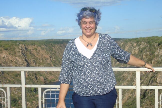 Foto de plano médio de uma mulher branca de cabelos curtos e tingidos em um tom de azul-violeta, que sorri para a câmera. Ela usa óculos escuros sobre a cabeça, uma blusa azul com estampas circulares brancas e gola em "V" branca, calças escuras e um colar com pingentes metálicos. Ela está em pé na frente de uma grade metálica cinza, que serve como parapeito para um mirante. Ao fundo, estende-se uma paisagem natural imponente, composta por cânions de rocha clara e vegetação rasteira, com o curso de um rio azul ao fundo do vale, sob um céu claro com poucas nuvens