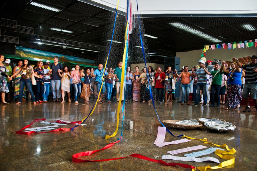 Esta fotografia registra um momento de celebração e mística durante um encontro de agricultores experimentadores. No centro de um amplo salão de piso brilhante, uma instalação simbólica se destaca: uma rede de pesca aberta em formato de cone, sustentada por fitas coloridas (azuis e amarelas) que descem do teto até o chão, onde se espalham fitas vermelhas e papéis com mensagens escritas. Ao redor dessa estrutura central, dezenas de homens e mulheres formam uma grande ciranda, de mãos dadas, simbolizando união e cooperação. Os participantes vestem roupas casuais e bonés, e ao fundo à direita, veem-se bandeirinhas coloridas de festa junina, reforçando o caráter cultural e comunitário do evento. A cena transmite um forte sentido de identidade coletiva e a importância dos rituais simbólicos na luta pela agroecologia.