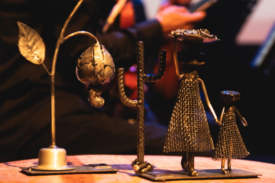 Esta fotografia em close-up apresenta pequenas esculturas feitas de metal reciclado, dispostas sobre uma superfície circular de madeira. À esquerda, destaca-se a representação de um caju pendendo de um galho com uma folha, fabricado com peças soldadas e polidas. Ao centro, há um cacto mandacaru feito de vergalhão texturizado. À direita, duas figuras humanas — uma maior e uma criança — são representadas com corpos de tela metálica e cabeças de parafusos, evocando a imagem de retirantes ou figuras regionais. O fundo está desfocado em tons quentes e escuros, revelando silhuetas de músicos e instrumentos, o que sugere que as peças fazem parte da decoração ou premiação do evento ocorrido no palco.