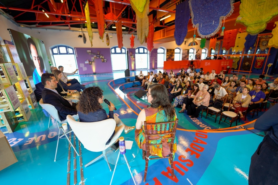 Esta fotografia colorida oferece uma perspectiva lateral e próxima do palco de um painel de discussão, capturando o evento sob o ponto de vista dos palestrantes. Em primeiro plano, três pessoas estão sentadas: à esquerda, um homem de terno escuro; ao centro, uma mulher de cabelos cacheados segura um microfone e fala para a plateia; à direita, vemos as costas de uma mulher vestindo uma blusa colorida em tons de verde, laranja e amarelo. Eles estão acomodados em poltronas brancas modernas sobre um piso azul brilhante, decorado com grandes frases em letras coloridas. No lado esquerdo, um grande telão exibe o rosto de uma participante que acompanha o evento remotamente. À direita, a plateia sentada em cadeiras de madeira preenche o salão, atenta à conversa. O teto alto é adornado com vigas vermelhas e inúmeros estandartes coloridos suspensos, incluindo peças em azul, amarelo e vermelho com franjas, que reforçam a identidade cultural e festiva do espaço.