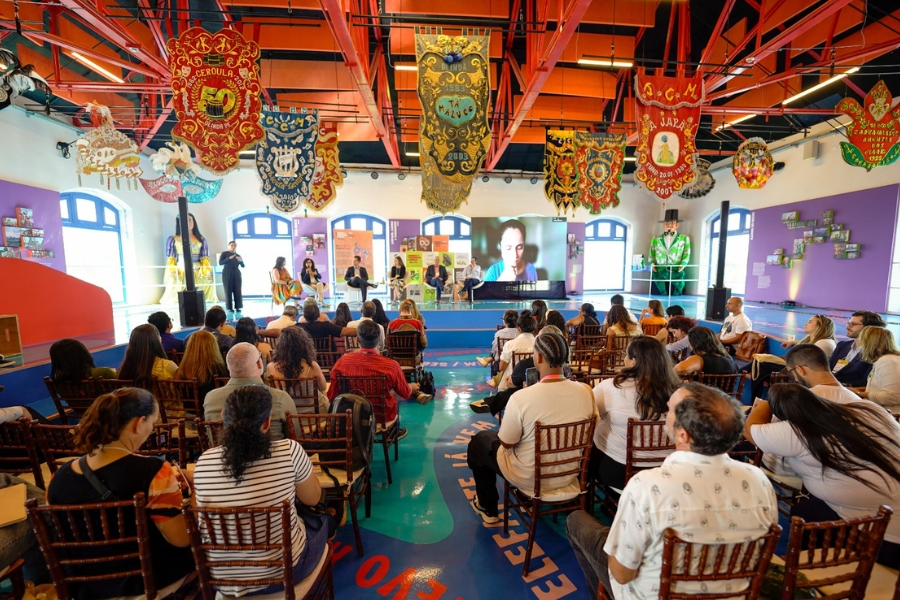 Uma fotografia colorida, capturada de um ângulo levemente elevado de trás da plateia, mostra uma palestra ou painel de discussão acontecendo em um salão vibrante e culturalmente rico, provavelmente no Paço do Frevo, em Recife. Cerca de quarenta pessoas estão sentadas em cadeiras de madeira voltadas para um palco baixo e curvo, onde seis palestrantes estão acomodados em poltronas brancas à frente de um telão que exibe uma pessoa em videoconferência. O ambiente possui um teto alto com vigas metálicas vermelhas, de onde pendem grandes e luxuosos estandartes de agremiações carnavalescas, ricamente bordados em tons de dourado, vermelho e azul. Ao fundo, grandes janelas em arco iluminam o espaço, ladeadas por bonecos gigantes de Olinda e exposições coloridas nas paredes. O chão é azul brilhante com inscrições em letras grandes e coloridas, completando a atmosfera festiva e institucional do evento.