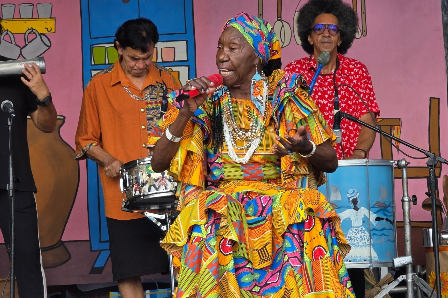 A foto registra uma apresentação cultural vibrante de uma mulher negra idosa de presença majestosa, que canta com um microfone vermelho na mão. Ela veste um traje exuberante e volumoso, com camadas de babados em tecidos africanos coloridos em tons de amarelo, laranja, azul e rosa, além de um turbante combinando e diversos colares de contas brancas e douradas. Ao seu redor, músicos a acompanham: à esquerda, um homem toca uma caixa de metal, e à direita, um homem de cabelos estilo black power e camisa vermelha toca um surdo azul decorado com a imagem de uma mulher negra. O cenário ao fundo é um painel lúdico e colorido que remete a uma cozinha rústica com utensílios domésticos desenhados
