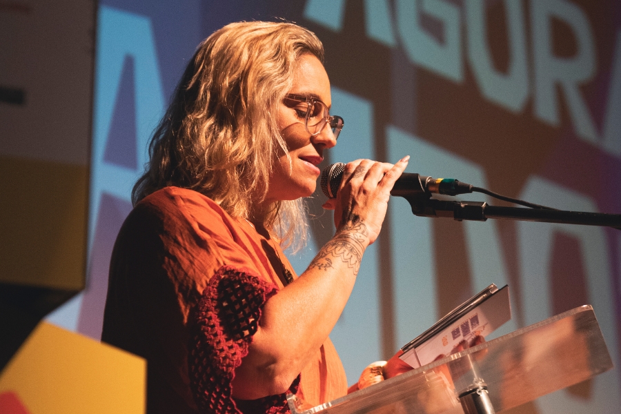 Esta é uma fotografia em plano médio e perfil de uma mulher de pele clara e cabelos loiros ondulados, falando ao microfone em um evento. Ela usa óculos de grau com armação clara e uma blusa em tom de terracota com detalhes em renda nas mangas. Com a mão direita, que possui uma tatuagem de estilo mandala no pulso, ela segura o microfone próximo à boca, enquanto a mão esquerda segura papéis sobre um púlpito de acrílico transparente. Ao fundo, uma projeção desfocada mostra letras grandes em tons de azul e vermelho, mantendo a identidade visual do evento anterior. A iluminação é quente e direcionada, destacando seu rosto e a expressão de quem realiza uma leitura ou discurso atento.