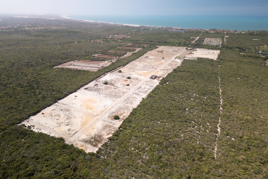 Fotografia aérea revela um grande loteamento em processo de terraplanagem, rasgando uma vasta área de vegetação nativa preservada. No centro, uma longa faixa retangular de solo arenoso e desmatado se estende diagonalmente pela imagem, dividida por uma estrada de terra central e marcações de lotes. Ao redor dessa clareira, o verde denso da vegetação litorânea domina a paisagem, interrompido apenas por outras pequenas áreas de construção ao fundo. Na linha do horizonte, o azul do mar encontra uma faixa de areia clara e áreas urbanizadas, sob um céu levemente brumoso