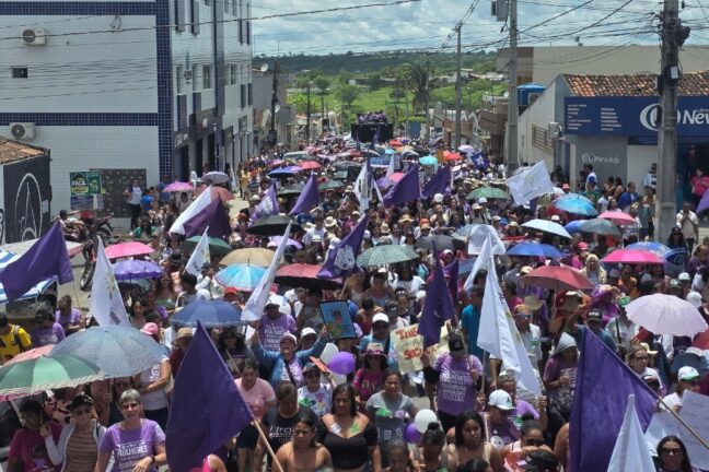 Uma multidão densa, composta majoritariamente por mulheres, caminha em direção à câmera, ocupando toda a extensão da via ladeada por prédios baixos e fiação elétrica. O grupo exibe uma identidade visual vibrante, com muitas participantes vestindo camisetas lilases e empunhando bandeiras roxas e brancas que flutuam sobre a massa. Para se protegerem do sol forte, diversas mulheres utilizam sombrinhas coloridas, criando um mosaico de cores que se mistura aos cartazes de reivindicação. Ao fundo, um trio elétrico. Tudo sob um céu claro com poucas nuvens