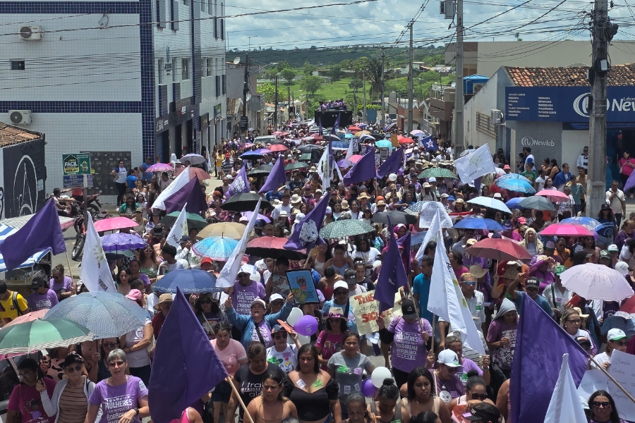 Uma multidão densa, composta majoritariamente por mulheres, caminha em direção à câmera, ocupando toda a extensão da via ladeada por prédios baixos e fiação elétrica. O grupo exibe uma identidade visual vibrante, com muitas participantes vestindo camisetas lilases e empunhando bandeiras roxas e brancas que flutuam sobre a massa. Para se protegerem do sol forte, diversas mulheres utilizam sombrinhas coloridas, criando um mosaico de cores que se mistura aos cartazes de reivindicação. Ao fundo, um trio elétrico. Tudo sob um céu claro com poucas nuvens