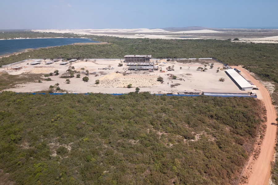 Esta fotografia aérea captura uma ampla área de construção em meio a uma paisagem natural preservada. No centro da imagem, destaca-se um terreno retangular de areia clara cercado por uma vegetação rasteira e densa, onde se vê a estrutura de concreto de um edifício principal de quatro andares em obras, além de outras fundações menores ao lado. À esquerda do canteiro, há uma lagoa de águas azuis margeada por verde, e, ao fundo, uma vasta linha de dunas de areia branca sob um céu azul limpo. Uma estrada de terra avermelhada contorna o lado direito do empreendimento, conectando a construção ao restante da região litorânea isolada