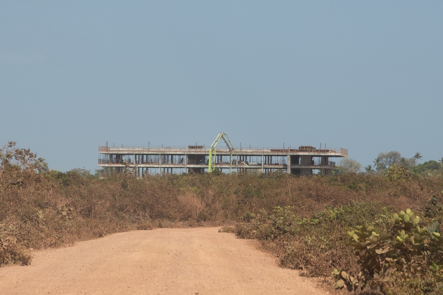 Esta fotografia horizontal, registrada ao nível do solo, mostra o esqueleto de concreto em construção. No primeiro plano, uma estrada de terra batida e clara se estende em direção ao centro da imagem, ladeada por uma vegetação de restinga baixa e predominantemente seca, em tons de marrom e verde opaco. Ao fundo, a estrutura retangular do edifício se destaca na linha do horizonte, exibindo dois pavimentos erguidos com colunas aparentes e uma bomba de concreto de braço longo e cor verde limão posicionada no topo. O céu é de um azul pálido e uniforme, sem nuvens