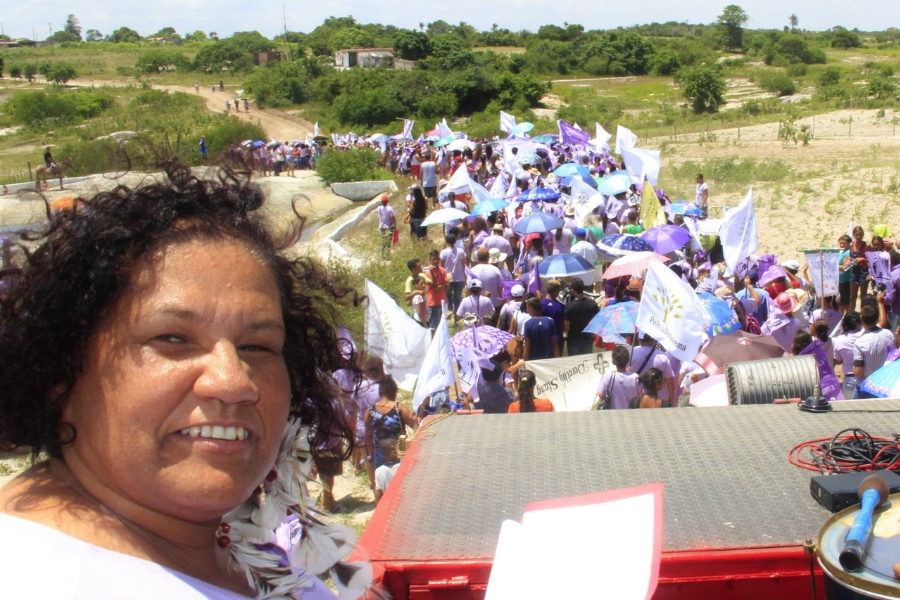 A imagem mostra, em primeiro plano à esquerda, o rosto sorridente de uma mulher negra de cabelos cacheados e escuros, vestindo uma blusa clara e brincos de penas brancas. Ela está posicionada no que parece ser a parte superior de um carro de som vermelho, onde se veem papéis e equipamentos de áudio. Ao fundo, sob a luz do dia, uma longa multidão caminha por uma estrada de terra em uma área rural com vegetação rasteira e árvores esparsas. Muitas pessoas vestem camisetas lilás e carregam bandeiras brancas e roxas, além de guarda-chuvas coloridos para se protegerem do sol, criando uma linha sinuosa que se estende até o horizonte.