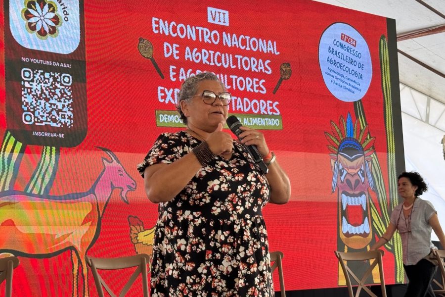 A imagem mostra mulher de pé em um palco, falando ao microfone durante um evento formal. Ela é uma mulher negra de pele clara, com cabelos curtos e grisalhos, usa óculos de grau redondos, um vestido preto com estampa floral branca e vermelha, e um bracelete largo no pulso direito. Ao fundo, um grande painel digital vermelho exibe o título "VII Encontro Nacional de Agricultoras e Agricultores Experimentadores", decorado com ilustrações coloridas de um bode, um cacto e uma figura mítica indígena, além de logotipos do Congresso Brasileiro de Agroecologia e um QR Code. Roselita segura o microfone com a mão esquerda enquanto gesticula com a direita, apresentando uma expressão serena e comunicativa, em um ambiente iluminado por uma estrutura de tenda branca ao fundo