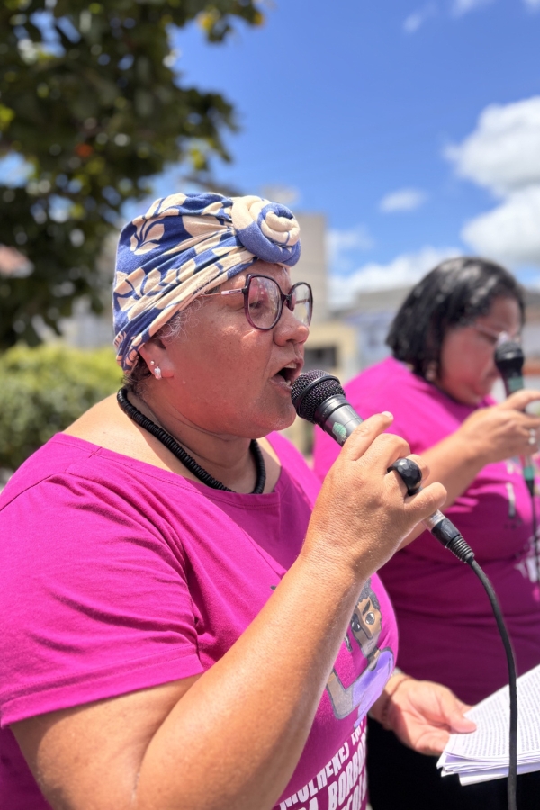 A imagem apresenta Roselita Vitor em plano médio, vista de perfil, enquanto discursa com vigor em um microfone preto. Ela é uma mulher negra de pele clara, usa óculos de grau de armação escura, um colar de contas pretas e um turbante azul com estampas florais bege, finalizado com um nó central. Veste uma camiseta de cor fúcsia vibrante que traz a ilustração de uma mulher e dizeres na parte inferior. Com a mão esquerda, ela segura algumas folhas de papel, possivelmente um manifesto ou roteiro. Ao fundo, uma segunda mulher com vestimenta semelhante também segura um microfone, indicando uma ação conjunta. O cenário é ao ar livre, com árvores e um céu azul com nuvens brancas, sugerindo um evento de mobilização social ou política.