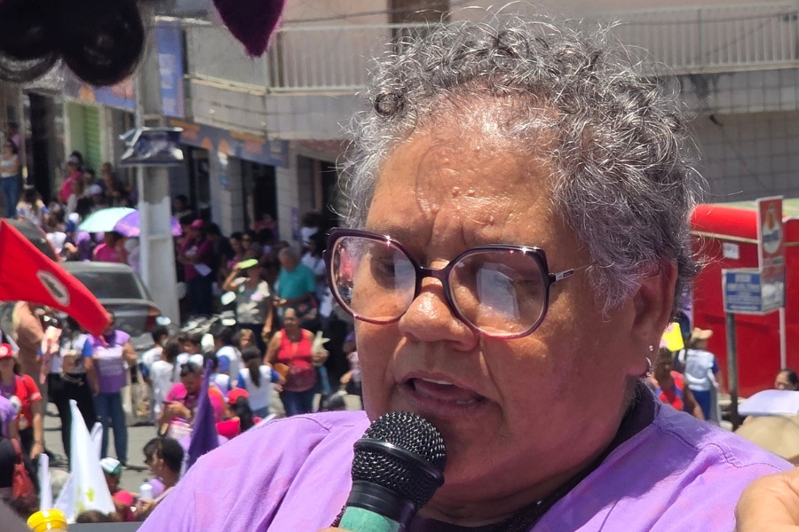 A foto em close de mulher de pele madura, traços negros e cabelos curtos e grisalhos levemente cacheados. Ela usa óculos de armação escura e fala ao microfone, que segura próximo à boca com uma expressão de seriedade e foco, veste uma camiseta de tom lilás, cor símbolo. O fundo da imagem está levemente desfocado, revelando a rua movimentada com diversas pessoas, prédios urbanos e uma bandeira vermelha à esquerda, indicando que ela está discursando durante movimento sob a luz direta do sol