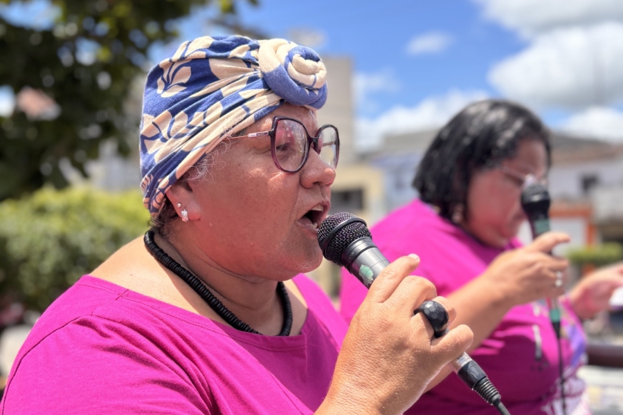 A imagem apresenta um plano médio e de perfil de uma mulher negra de pele clara, identificada como Roselita Vitor, falando ou cantando com entusiasmo em um microfone preto que segura com a mão direita. Ela tem uma expressão decidida, com a boca aberta, e usa óculos de armação escura, um colar de contas pretas e um turbante azul com estampas de flores bege, amarrado com um nó frontal. Roselita veste uma camiseta de cor fúcsia vibrante. Ao fundo, levemente desfocada, aparece outra mulher com vestimentas de cores semelhantes, também segurando um microfone, sugerindo um evento público ou manifestação ao ar livre sob um céu azul com nuvens esparsas e árvores ao redor