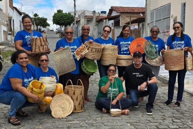 Esta fotografia captura um grupo de artesãos, majoritariamente mulheres, posando orgulhosamente em uma rua de paralelepípedos com suas criações manuais. A maioria veste camisetas azuis com a frase "Artesanato nada substitui o Digital", destacando a valorização do trabalho feito à mão. Eles exibem uma variedade de peças de cestaria e tecelagem, como cestos de diferentes tamanhos, bolsas, tapetes e descansos de prato, todos confeccionados em fibras naturais com detalhes em tons de palha, verde e laranja. Ao centro, um homem com camiseta preta e uma mulher com camiseta verde completam o grupo, que transparece um forte sentido de comunidade e preservação cultural local.