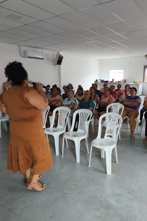 A imagem registra uma palestra ou reunião comunitária em um espaço amplo e iluminado, onde uma mulher negra, de costas para a câmera e vestindo um vestido longo na cor terracota, fala ao microfone para uma plateia atenta. O público é composto majoritariamente por mulheres, que estão sentadas em cadeiras de plástico brancas organizadas em fileiras sobre um piso de cimento queimado. Ao fundo, o ambiente possui paredes brancas com um aparelho de ar-condicionado instalado, e em um balcão lateral é possível observar garrafas e utensílios, sugerindo um momento de recepção ou intervalo. A cena transmite um tom de engajamento social e diálogo coletivo em um ambiente simples e funcional.