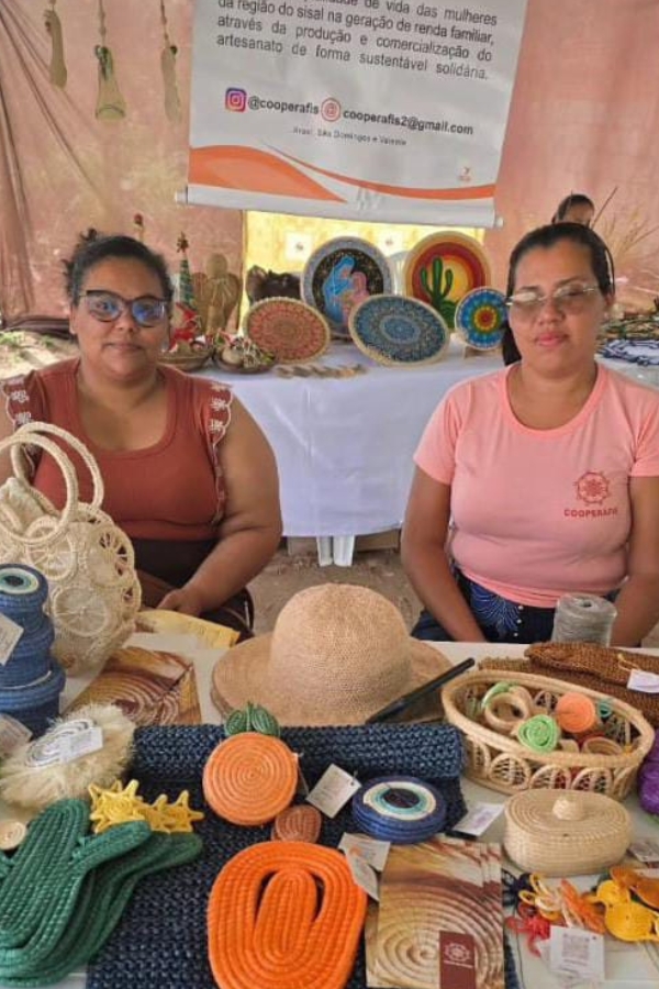 Nesta imagem, duas mulheres aparecem sentadas atrás de uma banca repleta de itens de artesanato em uma feira ou exposição. A mulher à esquerda usa óculos e uma blusa na cor terracota, enquanto a mulher à direita veste uma camiseta rosa com o logotipo "COOPERAFIS". Sobre a mesa, destaca-se uma variedade de produtos feitos de fibra natural (provavelmente sisal), incluindo chapéus, bolsas, cestos, descansos de prato coloridos e mandalas decorativas com motivos nordestinos, como cactos. Acima delas, um banner institucional detalha a missão da cooperativa de gerar renda para mulheres da região do sisal de forma sustentável e solidária, incluindo contatos de redes sociais. A cena reforça o protagonismo feminino na economia criativa e no fortalecimento das comunidades locais.