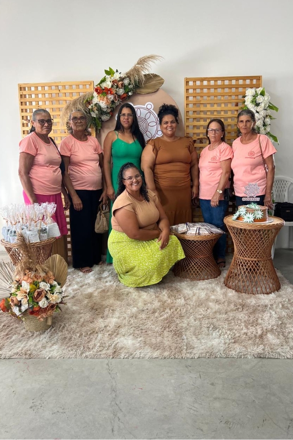 Esta fotografia registra um grupo de sete mulheres em um ambiente decorado para uma celebração ou evento institucional. Elas estão posicionadas à frente de um painel de madeira ripada adornado com flores e um logotipo circular, sobre um tapete felpudo bege. Quatro das mulheres vestem camisetas rosa idênticas com o logotipo da cooperativa, enquanto as outras três usam vestidos em tons de verde e terracota, incluindo uma mulher agachada ao centro com uma saia verde vibrante. O cenário é composto por móveis de fibra natural, como mesinhas que sustentam lembrancinhas embaladas e um bolo decorado com flores, reforçando a identidade artesanal e o clima de união do grupo.
