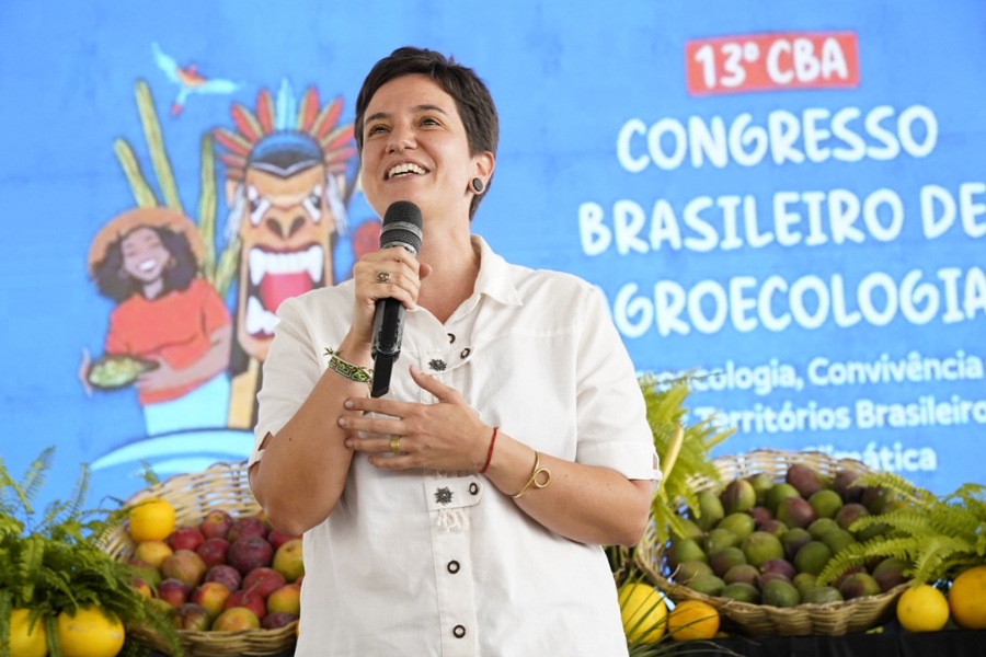 Uma mulher de pele clara e cabelos castanhos curtos sorri com entusiasmo enquanto fala ao microfone em um evento. Ela veste uma camisa branca de mangas curtas com botões escuros e detalhes bordados, além de acessórios como um anel dourado, pulseiras coloridas e um bracelete fino. Ao fundo, um painel azul vibrante exibe ilustrações coloridas de figuras humanas e a inscrição "13º CBA - Congresso Brasileiro de Agroecologia". À frente dela, compondo o cenário, há cestas de palha repletas de frutas diversas e folhagens verdes