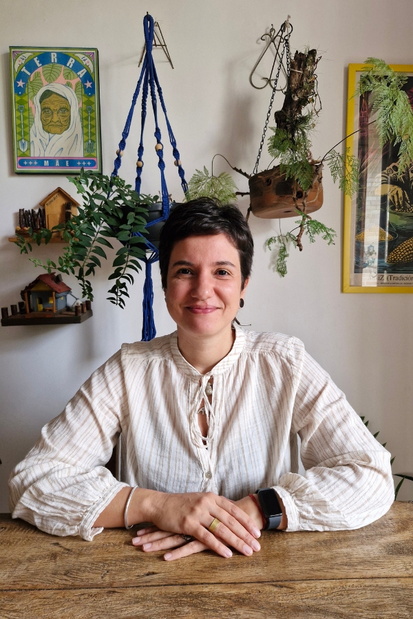 Uma mulher de pele clara, com cabelos castanhos curtos e um sorriso gentil, está sentada atrás de uma mesa de madeira rústica, com as mãos cruzadas sobre o tampo. Ela veste uma batinha bege clara com listras finas verticais, gola padre e um detalhe de amarração no decote. Em seus pulsos e dedos, nota-se uma pulseira prateada, um anel dourado e um relógio digital preto. O fundo é uma parede branca decorada com elementos afetivos e naturais: à esquerda, um quadro colorido com a ilustração de uma mulher idosa e a palavra "TERRA"; ao centro, um suporte de macramê azul com uma planta pendente e um tronco com folhagens; e à direita, um quadro em moldura amarela. Pequenas prateleiras de madeira com miniaturas de casas completam a composição