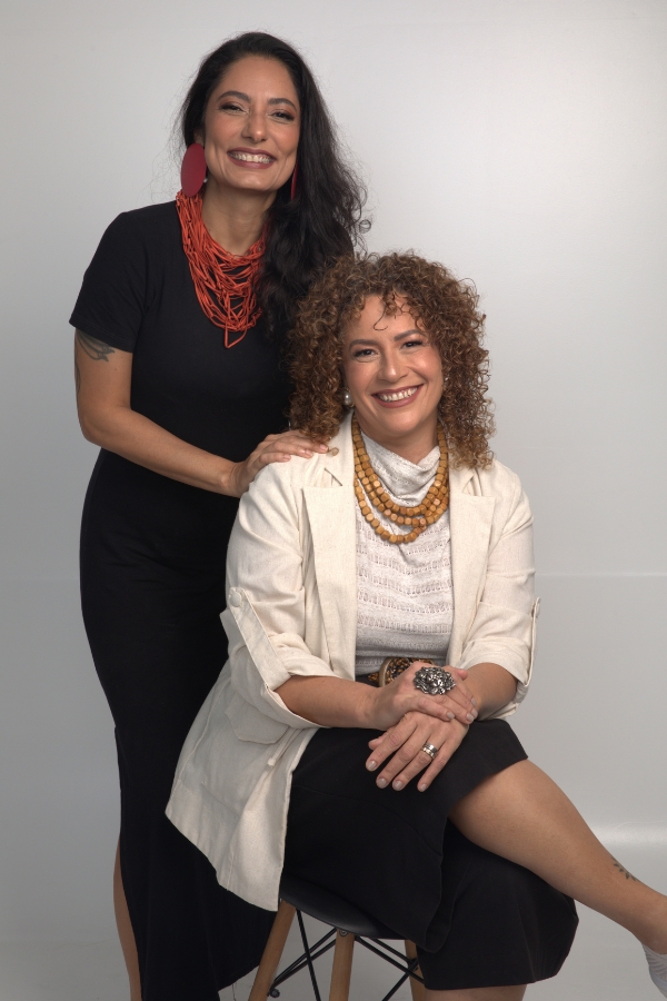 Esta fotografia colorida mostra duas mulheres posando sorridentes em um estúdio com fundo branco infinito. A da esquerda está de pé, com cabelos longos, escuros e ondulados; ela veste um vestido preto justo, brincos circulares grandes e vermelhos, e um colar de fios múltiplos em tom alaranjado, mantendo a mão direita apoiada no ombro da outra. Sentada à direita, a outra mulher exibe cabelos castanhos claros, curtos e cacheados; ela usa uma blusa texturizada off white sob um blazer leve de tom creme, acompanhados por um colar de contas de madeira em três camadas e um anel de destaque em formato de flor