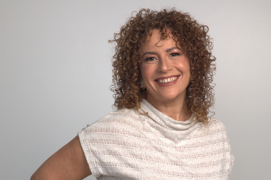 Esta foto colorida de estúdio captura uma mulher de meia-idade com pele morena clara, em pé contra um fundo cinza claro. Ela tem olhos castanhos expressivos e o rosto emoldurado por cabelos castanhos claros cacheados e volumosos, que caem logo acima dos ombros. Ela sorri calorosamente, olhando diretamente para a câmera, os dentes à mostra. Ela veste uma blusa off white de manga curta com gola careca, com uma textura sutil e listrada. Seus braços estão posicionados ao lado do corpo, um deles visível e ligeiramente dobrado, e ela usa um pequeno brinco redondo. Ela está posicionada no centro do enquadramento, preenchendo a maior parte da imagem