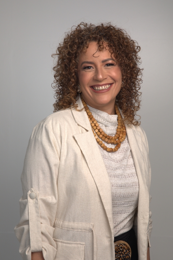 Nesta fotografia de estúdio em plano médio, uma mulher aparece sorridente contra um fundo cinza claro e neutro. De pele clara e rosto oval, ela possui cabelos castanhos claros, volumosos e bem cacheados, que emolduram seu rosto até a altura dos ombros. Ela veste um blazer de linho na cor creme com as mangas dobradas, sobreposto a uma blusa off-white de gola alta levemente texturizada. Destacam-se seus acessórios: um colar de três camadas feito de contas quadradas de madeira em tom caramelo e um cinto com uma fivela artesanal circular