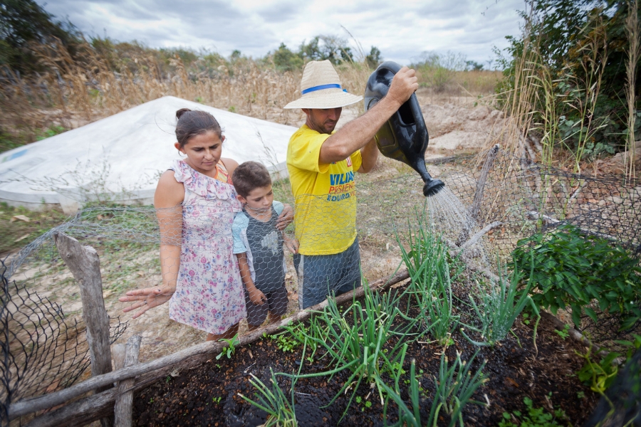 Nesta fotografia ao ar livre, uma família composta por um homem, uma mulher e uma criança pequena cuida de uma horta cercada em uma área rural. O homem, à direita, usa um chapéu de palha com fita azul e uma camiseta amarela, segurando um regador preto de plástico para molhar as fileiras de cebolinhas verdes que crescem no solo escuro. Ao seu lado, a mulher veste um vestido claro com estampa floral e observa a plantação com uma expressão serena, enquanto a criança, um menino, está entre os adultos, olhando atentamente para a água que cai. A horta é protegida por uma cerca de tela de arame e estacas de madeira. Ao fundo, destaca-se uma cisterna de placas branca, típica de regiões semiáridas, cercada por uma vegetação rasteira e seca sob um céu nublado