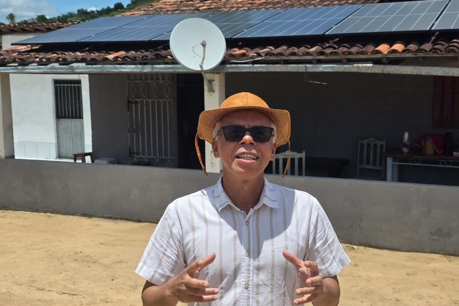Um homem de pele clara e cabelos grisalhos está em primeiro plano, centralizado, em um ambiente externo ensolarado. Ele usa óculos de sol escuros, uma camisa de botões branca com listras verticais finas e um chapéu de couro marrom de estilo regional (típico do sertanejo). O homem está com a boca levemente aberta, como se estivesse falando, e gesticula com as duas mãos à frente do corpo. Ao fundo, vê-se uma casa simples com um terraço sombreado por um telhado de telhas de barro. Sobre o telhado, estão instaladas várias placas de energia solar e uma antena parabólica branca. O chão à frente é de terra batida clara e, ao longe, sob um céu azul, avista-se o topo de uma colina com vegetação verde