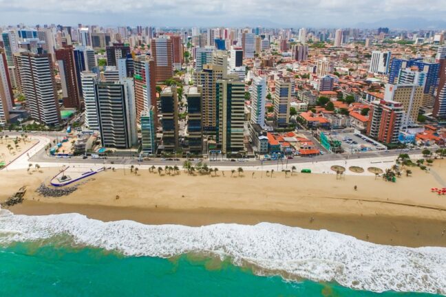Esta vista aérea captura a vibrante orla de Fortaleza, destacando o contraste entre a natureza e a urbanização densa da capital cearense. Em primeiro plano, as águas de um tom verde-esmeralda intenso quebram em espumas brancas sobre uma larga faixa de areia dourada, onde se alinham coqueiros e um calçadão moderno. Logo atrás da Avenida Beira-Mar, ergue-se um paredão de edifícios residenciais e comerciais de diferentes estilos arquitetônicos e alturas, que se estendem até o horizonte sob um céu levemente nublado. A imagem transmite a energia de uma metrópole litorânea tropical, equilibrando a serenidade do oceano com o dinamismo do cenário urbano.