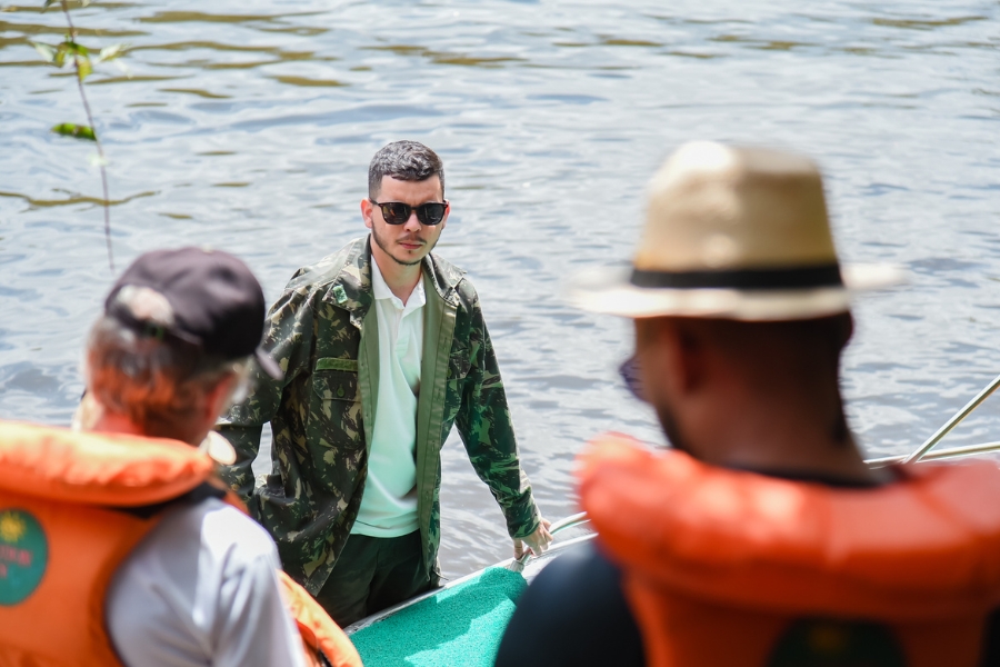 A imagem registra um momento de interação durante um passeio no Cânion do Rio Poti, focando na figura de um guia ou condutor local, conhecido como Jair do Cânion. Ele está posicionado em uma embarcação, vestindo uma jaqueta com estampa de camuflagem militar e óculos de sol, mantendo um olhar atento enquanto auxilia os visitantes. Em primeiro plano, as costas de dois turistas — um usando chapéu de palha e ambos com coletes salva-vidas alaranjados — criam uma moldura que direciona o olhar para o guia, evidenciando o aspecto humano e a segurança envolvida na exploração turística da região. Ao fundo, a superfície da água reflete suavemente a luz do dia, reforçando o cenário fluvial e a atmosfera de expedição guiada por esse importante personagem local.