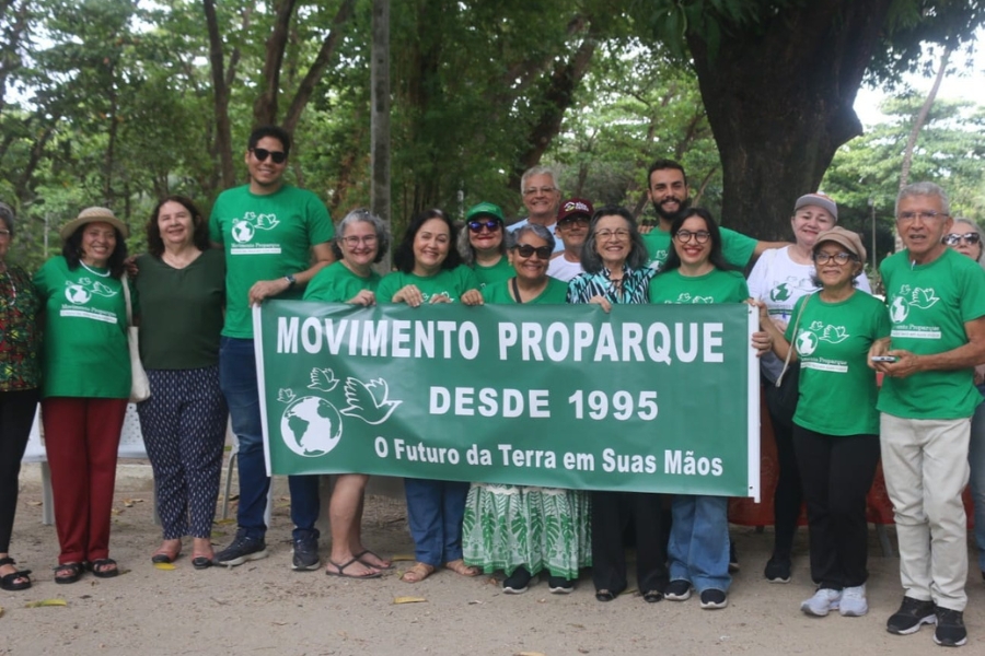 A imagem mostra um grupo de aproximadamente quinze pessoas reunidas em um ambiente arborizado, possivelmente um parque, posando para uma fotografia coletiva. O grupo, composto por homens e mulheres de diversas idades, exibe um banner verde central com letras brancas onde se lê "MOVIMENTO PROPARQUE - DESDE 1995" e o lema "O Futuro da Terra em Suas Mãos", acompanhado de ilustrações do globo terrestre e pombas brancas. Vários integrantes vestem camisetas verdes com a mesma logomarca do movimento, reforçando a identidade visual da causa ambientalista. O clima é de união e entusiasmo, com os participantes sorrindo para a câmera sob a sombra de grandes árvores, sobre um chão de terra batida e folhas secas.