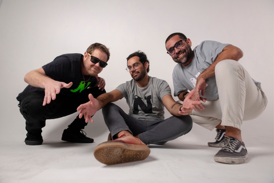 Esta fotografia promocional apresenta três homens em uma pose descontraída e dinâmica contra um fundo branco infinito, utilizando um ângulo de câmera baixo que enfatiza a proximidade com o espectador. À esquerda, um homem agachado veste camiseta preta com uma estampa verde e óculos de sol escuros; ao centro, um homem sentado de pernas cruzadas usa uma camiseta cinza com a letra "N" estampada e óculos de grau; e à direita, o terceiro homem, também agachado, veste uma camiseta cinza claro, calças brancas e óculos de armação escura, exibindo um sorriso aberto. Todos estendem as mãos em direção à lente em gestos acolhedores, criando uma composição triangular que transmite uma energia criativa, urbana e amigável