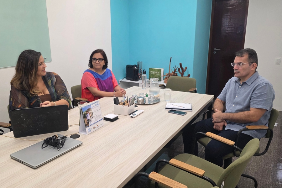 Três pessoas estão sentadas ao redor de uma mesa de reunião clara. Duas mulheres à esquerda e um homem à direita conversam em um ambiente profissional. Sobre a mesa, há um notebook, um calendário da Associação Caatinga e copos d'água, simbolizando um encontro estratégico.