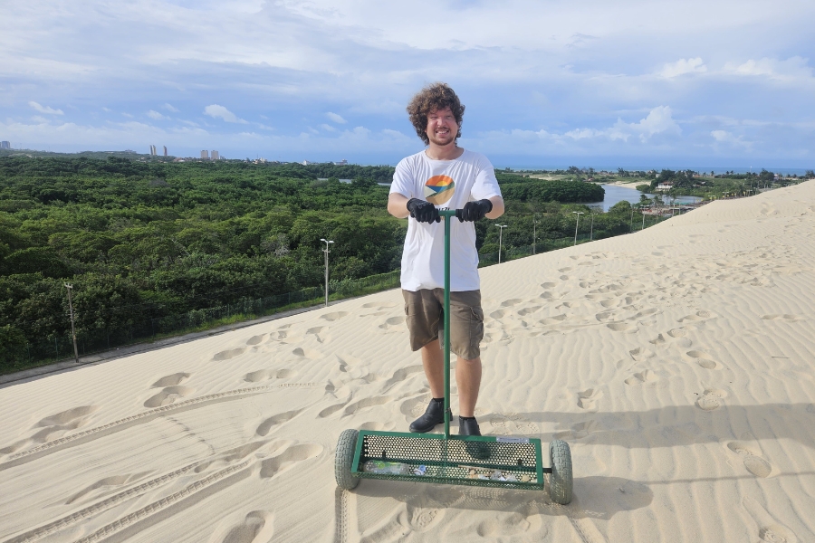 Esta fotografia ao ar livre registra um homem sorridente, de cabelos cacheados e volumosos, posando no topo de uma duna de areia clara enquanto segura um equipamento manual de limpeza. Ele veste uma camiseta branca com uma estampa circular colorida, bermuda cargo marrom, luvas pretas e botas escuras, mantendo as mãos no guidão de uma ferramenta verde composta por um rolo aramado que coleta resíduos da areia. O cenário ao fundo revela um contraste marcante entre a textura ondulada da duna, repleta de pegadas, e uma vasta área de vegetação densa e verdejante, cortada por um braço de rio ou lago sob um céu nublado, capturando uma ação de preservação ambiental em uma paisagem natural litorânea.