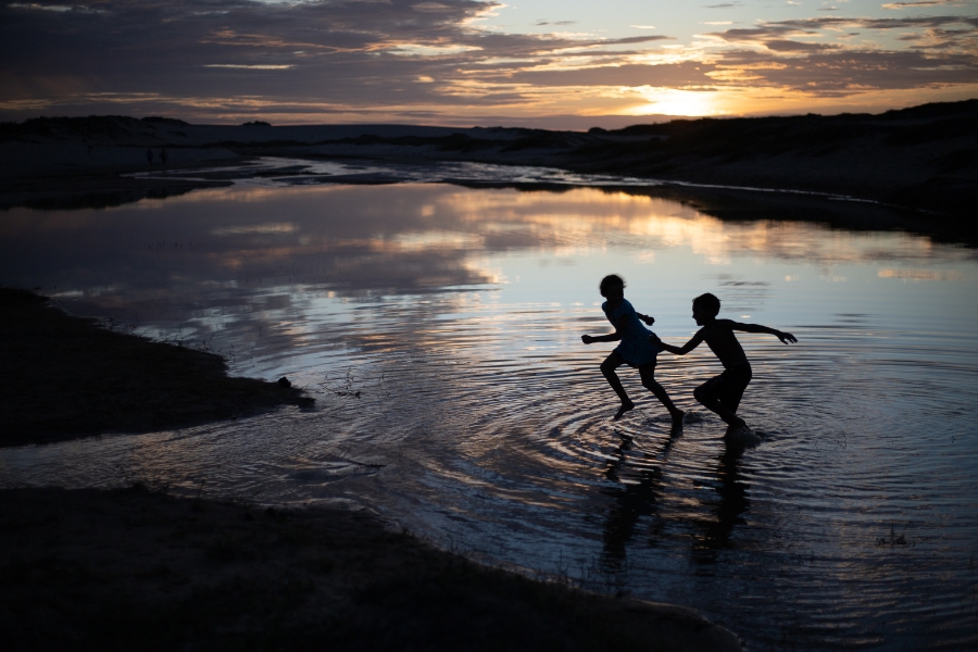 A fotografia captura um momento lúdico e poético de duas crianças em silhueta, correndo e brincando nas águas rasas de uma lagoa ou braço de mar durante o pôr do sol. O cenário é marcado por um céu dramático em tons de dourado, laranja e violeta, que se reflete de forma quase perfeita na superfície espelhada da água, criando uma continuidade visual entre o céu e a terra. Ao fundo, as dunas e a vegetação rasteira formam uma moldura escura contra o horizonte iluminado, enquanto o movimento das crianças gera ondulações suaves na água, reforçando uma atmosfera de liberdade, infância e integração com a natureza exuberante do território Tremembé.
