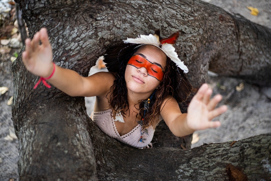 Esta imagem retrata uma jovem indígena Tremembé em um momento de profunda introspecção e reverência, posicionada entre os troncos robustos de uma árvore antiga. Ela veste um top de crochê e um cocar adornado com penas brancas, pretas e vermelhas, destacando-se pela marcante pintura facial em urucum que cobre a área dos olhos. Com os olhos suavemente fechados e o rosto voltado para cima, ela estende os braços em direção à câmera, criando uma perspectiva que convida o espectador para o seu espaço sagrado. A composição utiliza o enquadramento natural dos galhos texturizados para focar na expressão de serenidade da jovem, simbolizando uma conexão visceral entre a nova geração e a força da ancestralidade que emana da natureza.