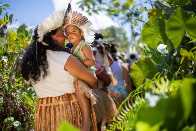 A imagem captura um momento afetuoso e geracional do povo Tremembé, onde uma mulher indígena carrega uma criança pequena no colo em meio a uma trilha cercada por vegetação tropical exuberante. Ambas utilizam cocares de penas claras e trajes tradicionais, como a saia de fibra natural, além de ostentarem a pintura facial vermelha de urucum cruzando as bochechas, simbolizando a continuidade da identidade cultural. Enquanto a mulher sorri com ternura, a criança olha diretamente para a câmera com uma expressão serena e curiosa, destacando-se sob a luz solar que filtra através das folhas. A composição, rica em tons de verde e iluminada pelo sol, transmite uma poderosa mensagem de preservação, cuidado materno e o florescimento das tradições ancestrais nas novas gerações.