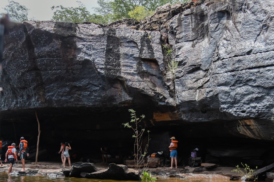 Foto de um grupo de turistas explorando uma imensa formação rochosa que avança sobre a margem do rio, criando uma espécie de abrigo ou gruta natural. Os visitantes aparecem equipados com coletes salva-vidas alaranjados, caminhando e descansando sob a sombra da imponente parede de pedra escura, que exibe texturas rugosas e marcas de erosão milenar. No topo da rocha, a vegetação nativa se debruça sobre a estrutura, enquanto no plano inferior, a proximidade com a água e a escala humana diante dos paredões enfatizam a magnitude geológica do local, revelando o cânion como um destino de aventura e contemplação